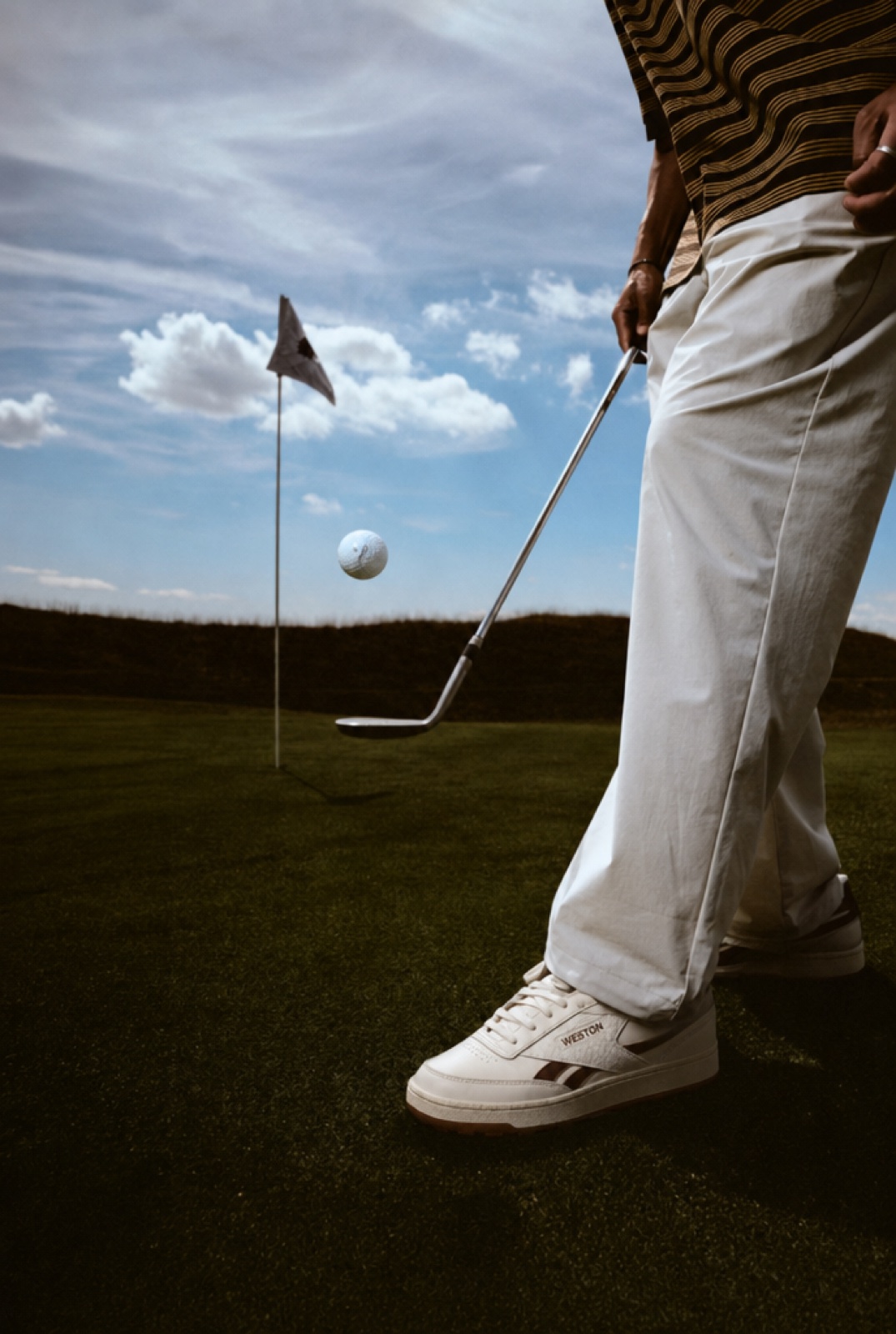 The Court Golf Shoe in Espresso, a golfer bouncing a ball on the club face beside the flag.